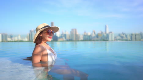 Young-rich-Asian-woman-relaxing-in-a-luxurious-infinity-pool-with-an-incredible-view-of-Bangkok-cityscape-in-the-background,-she-is-leaning-on-her-elbows,-side-view-slow-motion