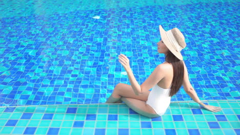 Young-American-Woman-Sitting-on-the-Swimming-Pool-Border-With-her-Legs-Inside-Water-and-Touching-Straw-Sunhat-Brim-with-Fingers,-Bora-Bora-hotel,-Slow-motion-elevated-view,-copy-space
