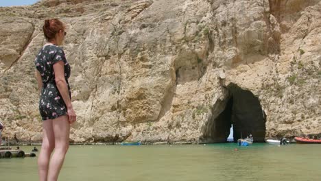 Girl-Standing-In-The-Shore-Looking-At-Sea-Cave-With-Boats-On-A-Sunny-Day-In-Inland-Sea,-Malta