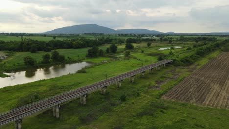 Aerial-footage-towards-a-beautiful-landscape-and-mountains-in-the-horizon-revealing-an-elevated-provincial-railway,-ponds,-trees,-and-farmlands,-Muak-Klek,-Saraburi,-Thailand