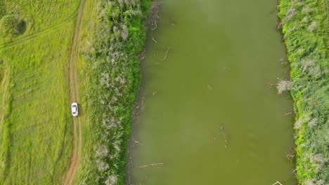 Aerial-footage-of-a-canal-proving-water-to-farms-while-a-car-is-revealed-moving-on-a-dirt-road,-Saraburi,-Thailand