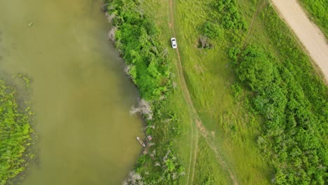 A-car-moving-on-a-dirt-road-going-to-the-main-road-next-to-the-river,-aerial-capture,-Muak-Klek,-Saraburi,-Thailand