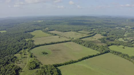 4K-aerial-high-over-green-fields-surrounded-by-woodland-with-hills-in-distance