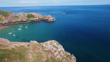 Drone-footage-of-the-turquoise-waters-of-a-bay-where-five-sailboats-are-moored-in-a-sunny-day-at-Sark-Island,-Channel-Islands