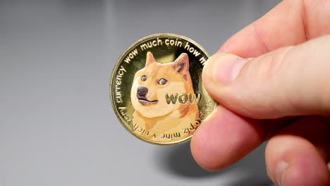 Goldene-Dogecoin-Münze,-Die-Zwischen-Den-Fingern-In-Der-Hand-Auf-Dem-Gey-Tisch-Gehalten-Wird