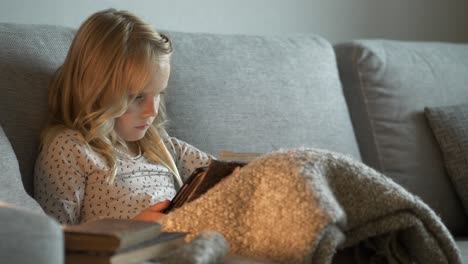 Young-blonde-girl-sitting-on-couch-with-focus-to-interesting-book,-handheld-view