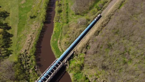 Aerial-track-shot-of-blue-train-passing-bridge-in-rural-area-of-Buenos-Aires-during-summer