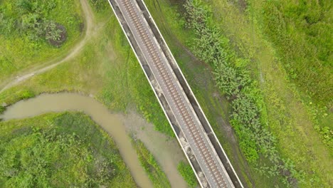 Aerial-ascending-footage-closer-from-the-elevated-provincial-railway-going-higher-revealing-a-wider-perspective-of-grassland-and-water,-Muak-Klek,-Saraburi,-Thailand