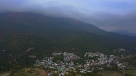 drone-shot-of-a-old-small-village-at-the-bottom-of-a-mountain-with-a-forest-during-the-day,-clouds-are-sticking-to-the-top-of-the-mountain