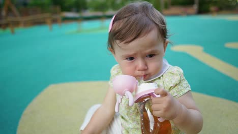 Portrait-of-1-year-old-cute-baby-drinking-juice-from-bottle-holding-it-with-one-hand-in-sitting-at-outdoor-Playground
