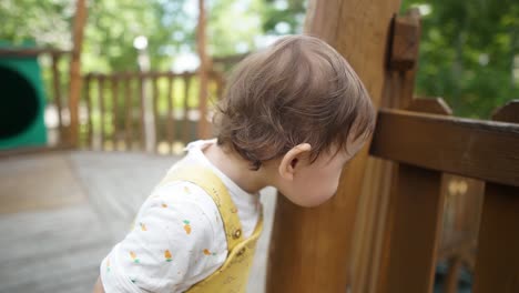 Adorable-little-baby-girl-lean-on-the-wooden-column-and-looking-throw-the-wooden-fence-out-of-curiosity-at-kid's-outdoor-playground-daytime,-slow-motion-close-up