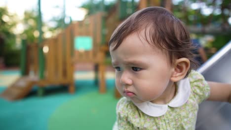 Unhappy-sad-or-worried-face-of-a-one-year-baby-girl-standing-on-the-slide-at-outdoor-kid's-playground-looking-seriously-at-something