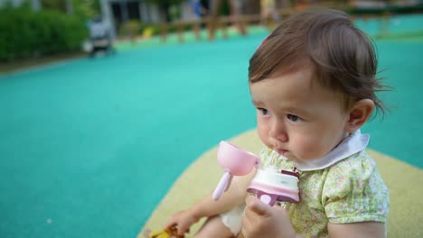 A-sweet-one-year-old-girl-is-sitting-on-playground-floor-holding-the-bottle-in-hands,-drinking-water,-template-slow-motion-copy-space