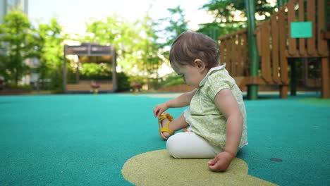 Lonely-One-year-old-cute-little-baby-girl-sitting-on-the-floor-of-outdoor-kid's-playground-alone-and-pick-up-small-stone-from-the-ground