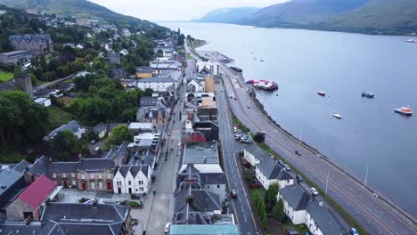Cars-Driving-Along-Road-Near-Loch-in-Fort-William-Town,-West-Highland-Museum,-Town-Centre---Aerial-Drone-4K-HD-Footage-Rise-Up