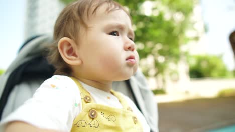 Little-1-year-baby-girl-staring-at-kids-in-the-playground-while-she-is-sitting-in-a-stroller-with-a-serious-face-expression,-close-up-in-slow-motion,-low-angle-summer-daytime