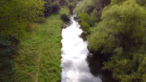 Aerial-flight-over-the-small-river-with-the-pathway-on-the-left-bank-and-cloud-reflections-on-the-water---Little-Ouse-near-Thetford-in-the-UK