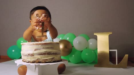 Süßes-Lateinamerikanisches-Kleinkind-Feiert-Sein-1.-Jahr-Und-Entfernt-Die-Nummer-Eins-Von-Seinem-Kuchen-Mit-Grünen-Luftballons-Im-Hintergrund