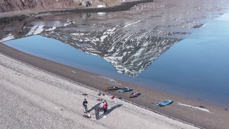 Group-of-kayaking-friends-on-shore-of-Holmanes,-reflection-of-Hólmatindur-mountain-on-flat-water-surface,-aerial