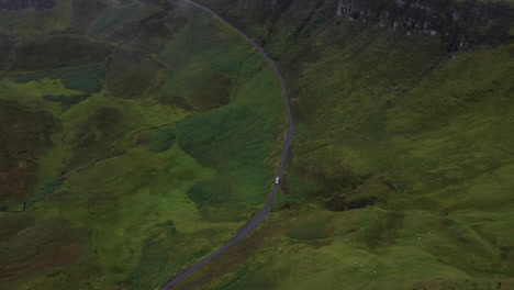 Cinematic-downward-angle-drone-shot-coming-through-the-clouds,-showing-a-vehicle-on-the-mountain-side-of-Quiraing-in-Scotland