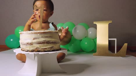 Süßes-Lateinisches-Kleinkind,-Das-Seinen-1.-Geburtstag-Feiert-Und-Im-Hintergrund-Einen-Kuchen-Mit-Grünen-Luftballons-Zerschmettert