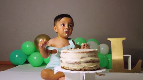 Süßes-Lateinisches-Kleinkind,-Das-Seinen-1.-Geburtstag-Feiert-Und-Einen-Kuchen-Mit-Grünen-Luftballons-Im-Hintergrund-Beißt-Und-Zerschmettert