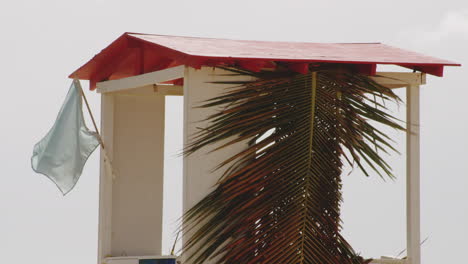 Flagge-Weht-Im-Wind-An-Der-Rettungsschwimmerhütte-Mit-Kokosnussblättern-Am-Balneario-Del-Escambron,-Strand-In-San-Juan,-Puerto-Rico