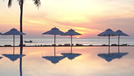 A-view-from-across-a-resort-swimming-pool-framed-by-beach-umbrellas,-a-lone-fishing-boast-travels-across-a-stunning-colorful-sunset