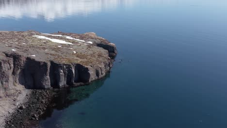 Rocky-cliff-shore-of-Eskifjörður-fjord-with-distant-kayakers-near-coast,-aerial