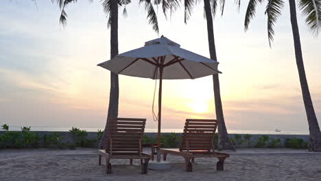 Close-up-of-two-empty-lounge-chairs-and-shade-umbrella-on-the-beach-as-the-light-from-the-sunset-fades