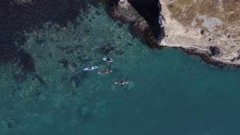 Above-kayakers-in-blue-clear-Iceland-water-near-rocky-shore-in-fjord,-aerial