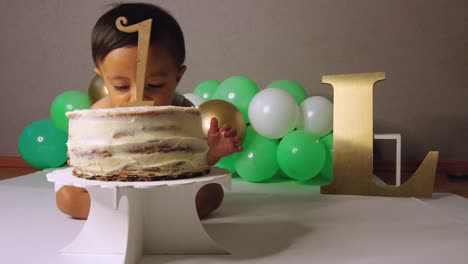 Cute-latin-baby-toddler-celebrating-his-birthday-biting-his-cake-and-smiling-with-green-balloons-in-the-background