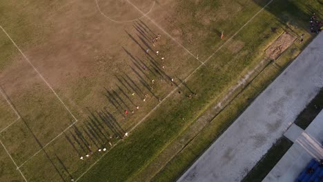 Aerial-birds-eye-shot-of-young-team-running-over-dried-grass-soccer-field-during-training-session-at-sunset