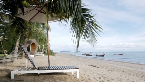 Beach-chairs-under-a-coconut-tree-on-the-beach-overlooking-the-ocean-and-boats-in-Satun,-Thailand