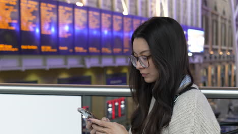 Young-woman-texting-on-her-smartphone-while-waiting-for-her-train,-timetable-boards-can-be-seen-in-the-background