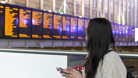 Young-woman-on-her-phone-checks-the-train-station-departure-board-before-leaving-to-catch-her-train