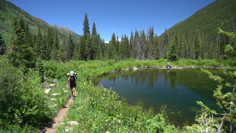 Junge-Weibliche-Wanderer-Mit-Rucksack,-Die-An-Einem-Sonnigen-Sommertag-Am-Teich-In-Der-Grünen-Landschaft-Der-Felsigen-Berge-Spazieren-Gehen,-Colorado-Usa