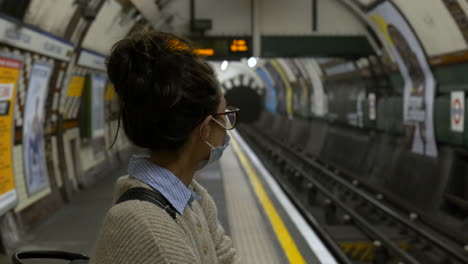 Woman-wearing-a-face-mask-waits-in-an-empty-tube-station,-London,-UK