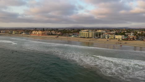 Imperial-Beach-With-Surfers-In-The-Ocean-And-People-Relaxing-On-The-Seashore-With-Waterfront-Buildings-In-The-Background