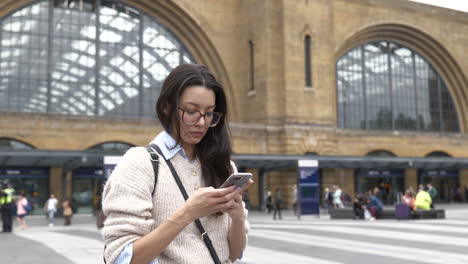 Attraktive-Junge-Frau-Mit-Ihrem-Smartphone,-Während-Sie-Vor-Dem-Haupteingang-Zum-Bahnhof-Kings-Cross-In-London-Wartete