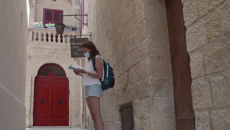 Female-Tourist-In-Facemask-Wandering-In-The-Steert-Of-Mdina-Looking-For-Destinatination-Through-Guidebook-In-Malta