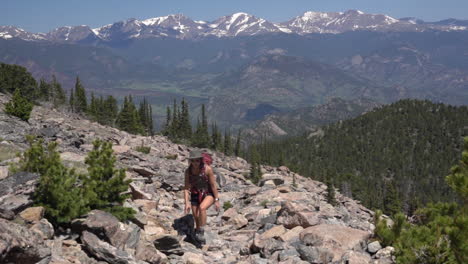 Young-Female-Hiker-Walking-Alone-on-Rocky-Mountain-Slopes-on-Sunny-Summer-Day-With-Snow-Capped-Summits-in-Skyline
