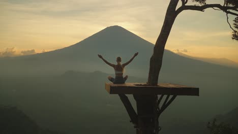 Woman-lifting-arms-on-platform-doing-yoga-in-front-of-majestic-Mount-Agung,-sunset