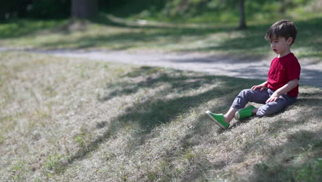 A-young-little-boy-is-sitting-on-a-green-field-under-dappled-light-and-throwing-rock