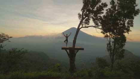 Woman-balancing-on-one-leg-doing-yoga-tree-pose-on-wood-platform,-Mount-Agung,-sunset