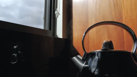 Wide-shot-of-black-household-domestic-kitchen-kettle-slowly-boils-for-cup-of-tea-coffee-with-steam-water-vapor-coming-out-of-metal-spout-with-window-in-background