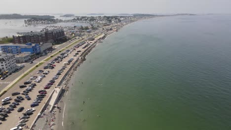 Birds-Flying-Over-Nantasket-Beach-In-Hull,-Massachusetts-With-People-Swimming-And-Cars-Parked-At-Summer