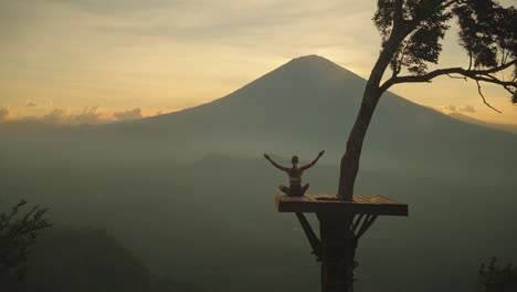 Woman-raising-arms-in-lotus-pose-on-scenic-wooden-platform-looking-at-Mount-Agung,-sunset
