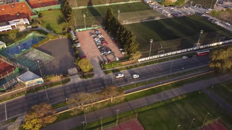 Aerial-tracking-shot-of-two-white-cars-driving-on-avenue-beside-soccer-field-of-Buenos-Aires-during-sunset