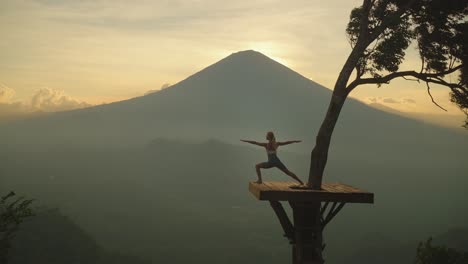 Woman-on-Balinese-platform-of-Lahangan-Sweet-doing-warrior-pose-with-view-of-volcano-Agung,-sunset